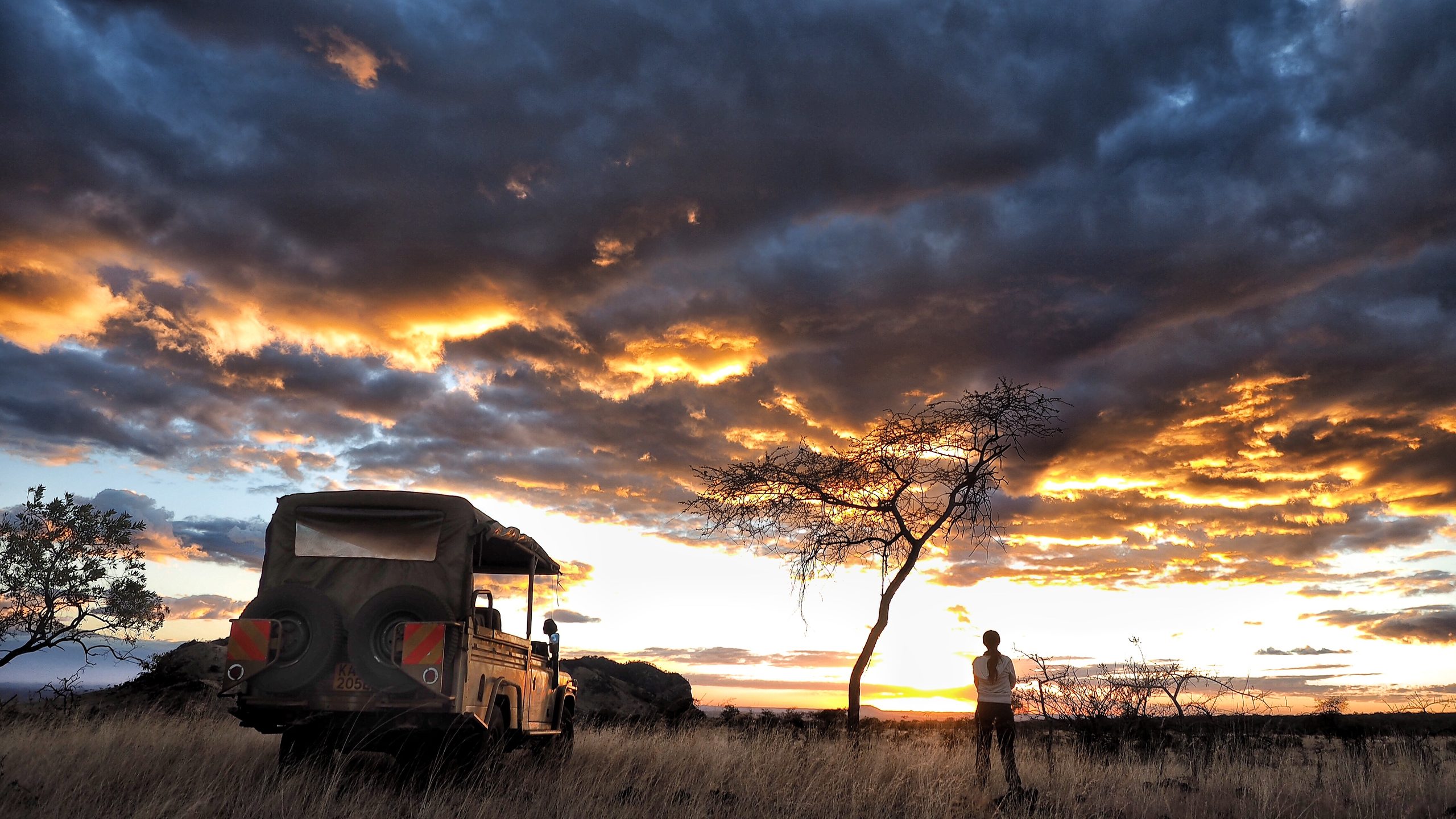 A safari vehicle and a lone figure silhouetted beneath an acacia tree against a dramatic cloud-filled sunset sky in the Chyulu Hills, Kenya